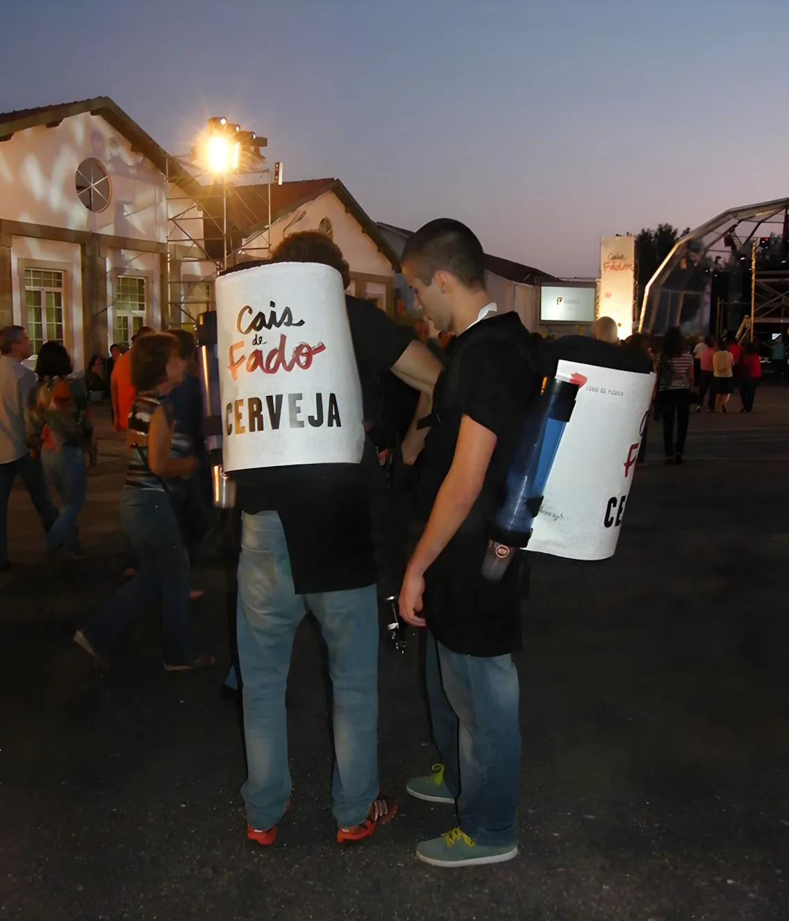 Duas pessoas com mochilas de bebida "Cais de Fado Cerveja" num evento ao anoitecer